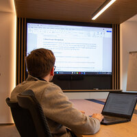 Person am Tisch in einem Besprechungsraum mit Laptop und vernetzten Fernseher
